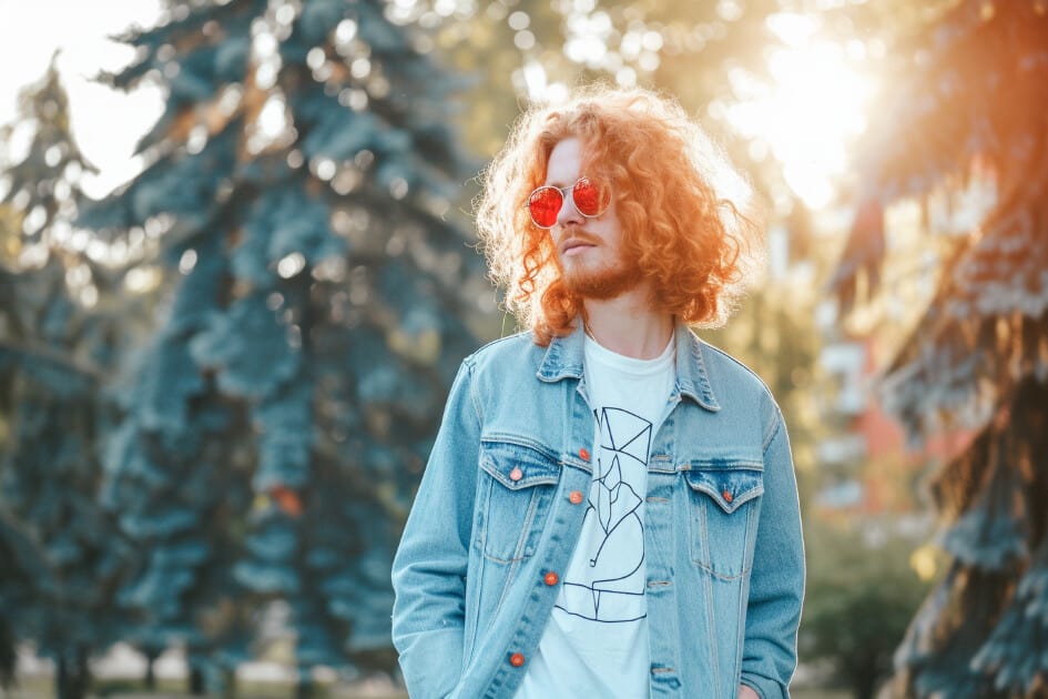 Casual outdoor fashion shot. Male, Caucasian ethnicity, curly red hair, playful expression. Vintage denim jacket, white graphic tee, red sunglasses. City park background, sunny day. Natural light, relaxed and cheerful mood.